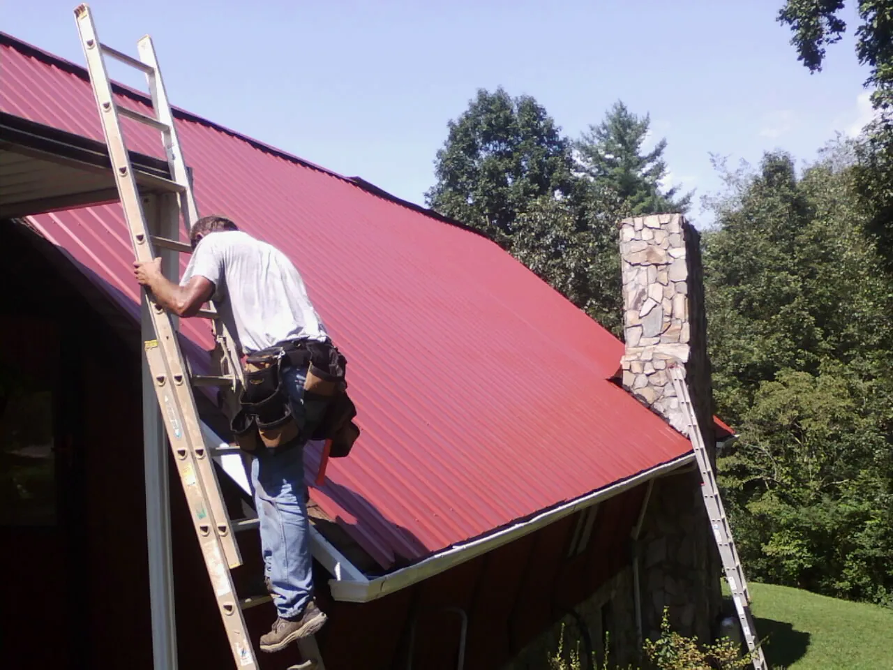Roofer on a ladder beside a red metal roof and stone chimney — Soffit & Fascia Repair by Roofing Brothers in Persia